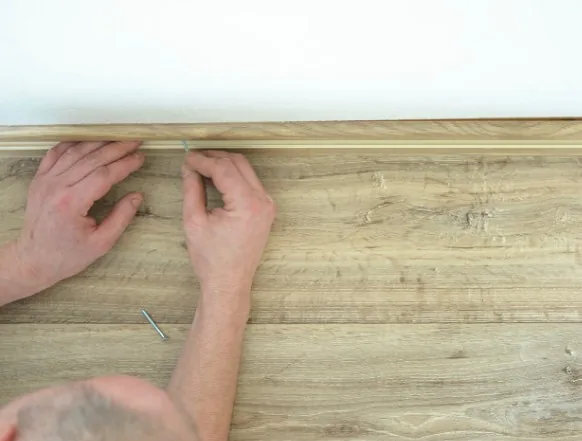 Manos clavando un rodapié de madera clara sobre suelo de tarima con clavo visible en Burgos