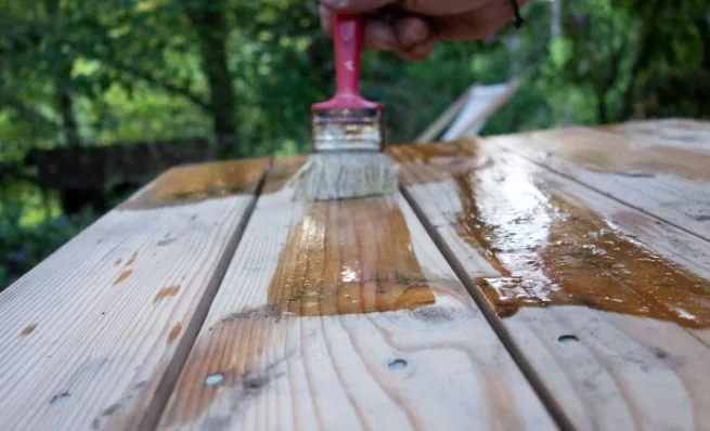 Mano aplicando barniz con brocha roja sobre mesa de madera de pino en exterior en Burgos