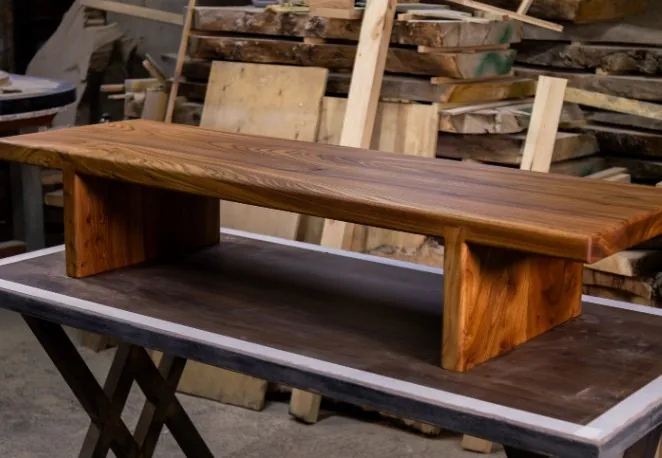 Banco/mesa baja de madera maciza con veta natural sobre patas macizas, en taller con tablones apilados al fondo en Burgos
